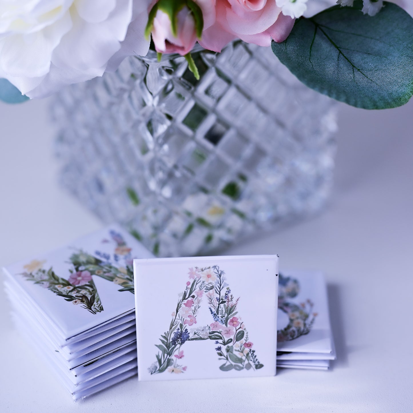 Stack of fridge magnets with floral design and letter 'A' on a white surface with flowers in the background