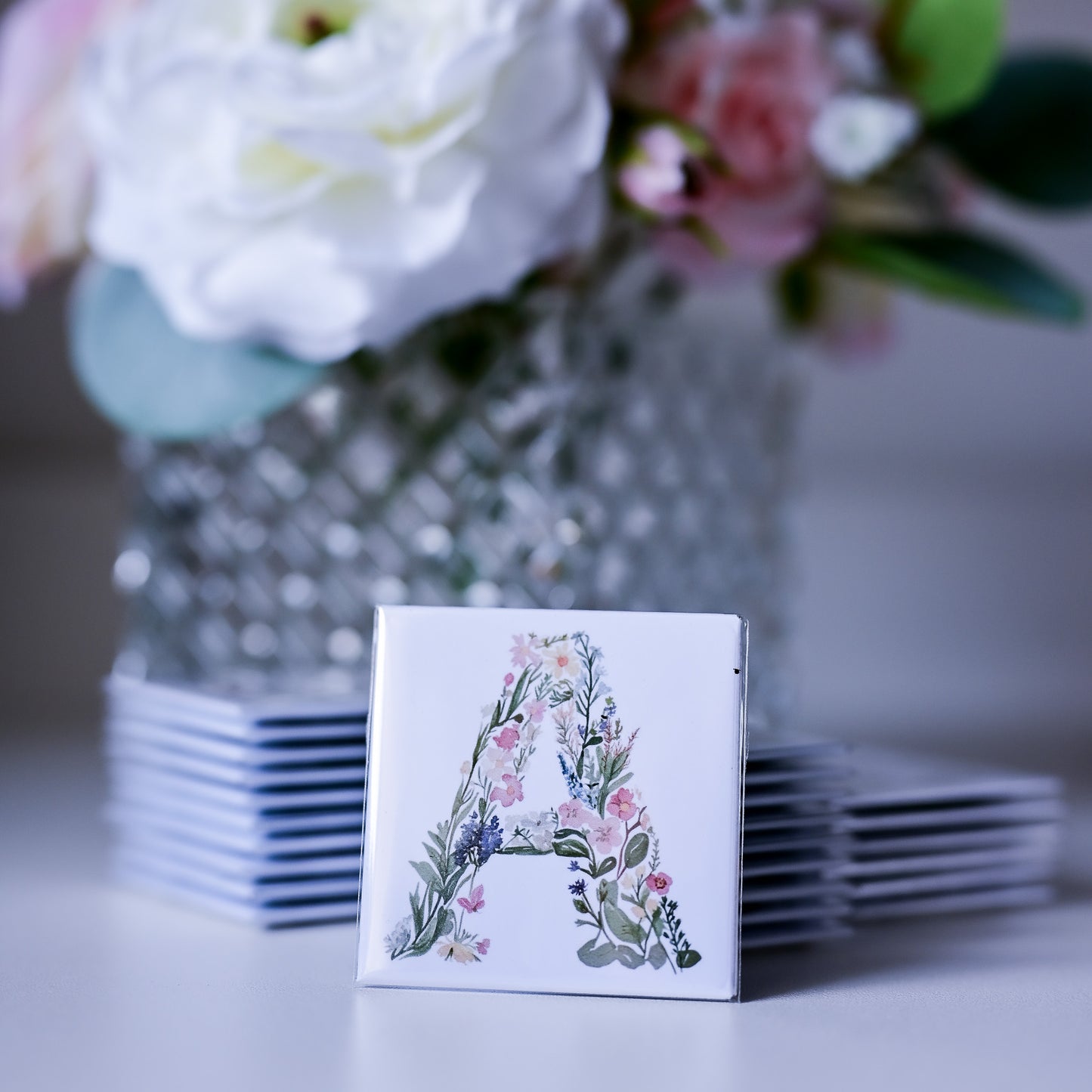Stack of fridge magnets with floral letter 'A' design in front of a vase with flowers.