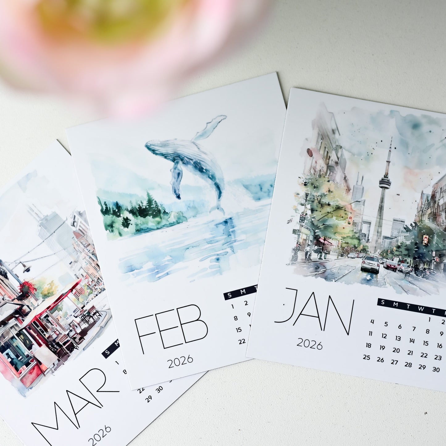 Photo Magnet Calendar sheets with watercolor designs of a whale, cityscape, and bus on a white surface.