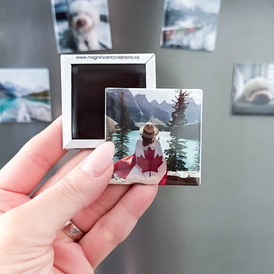 Hand holding a small photo magnet of a person with a Canadian flag draped over their shoulders, with more photos on a fridge in the background.