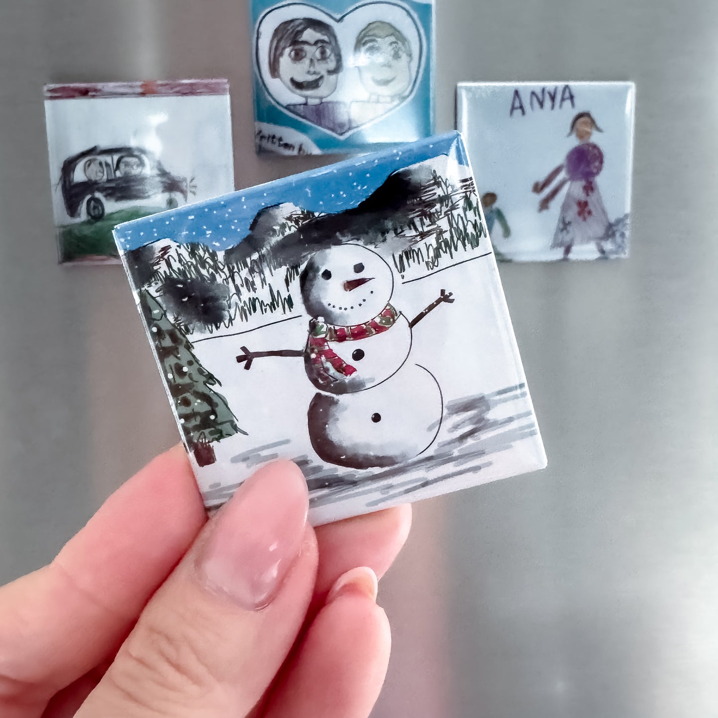 Hand holding a small photo magnet with a snowman illustration against fridge with other custom photo magnets displayed.