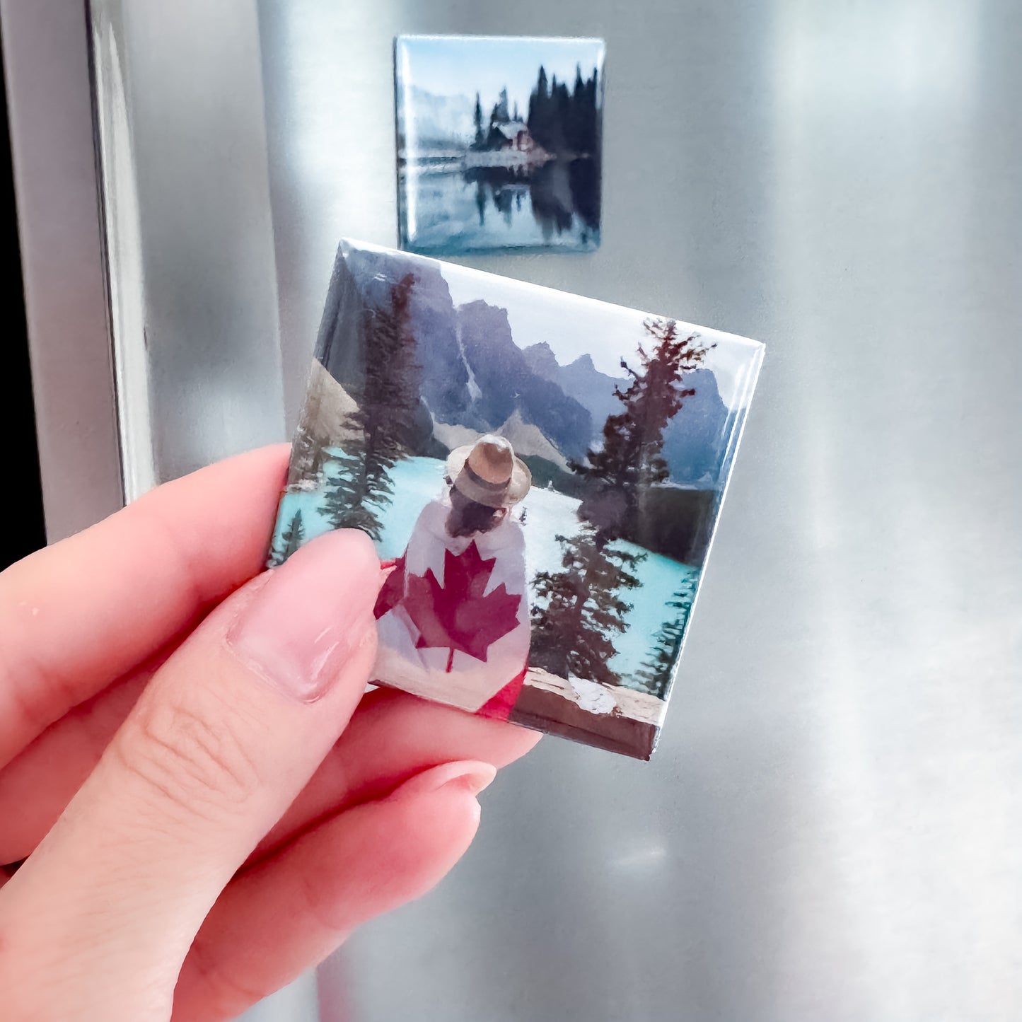 Hand holding a small photo magnet with a design of a lady with a Canadian flag against a mountain and lake background, with another similar magnet on a fridge.