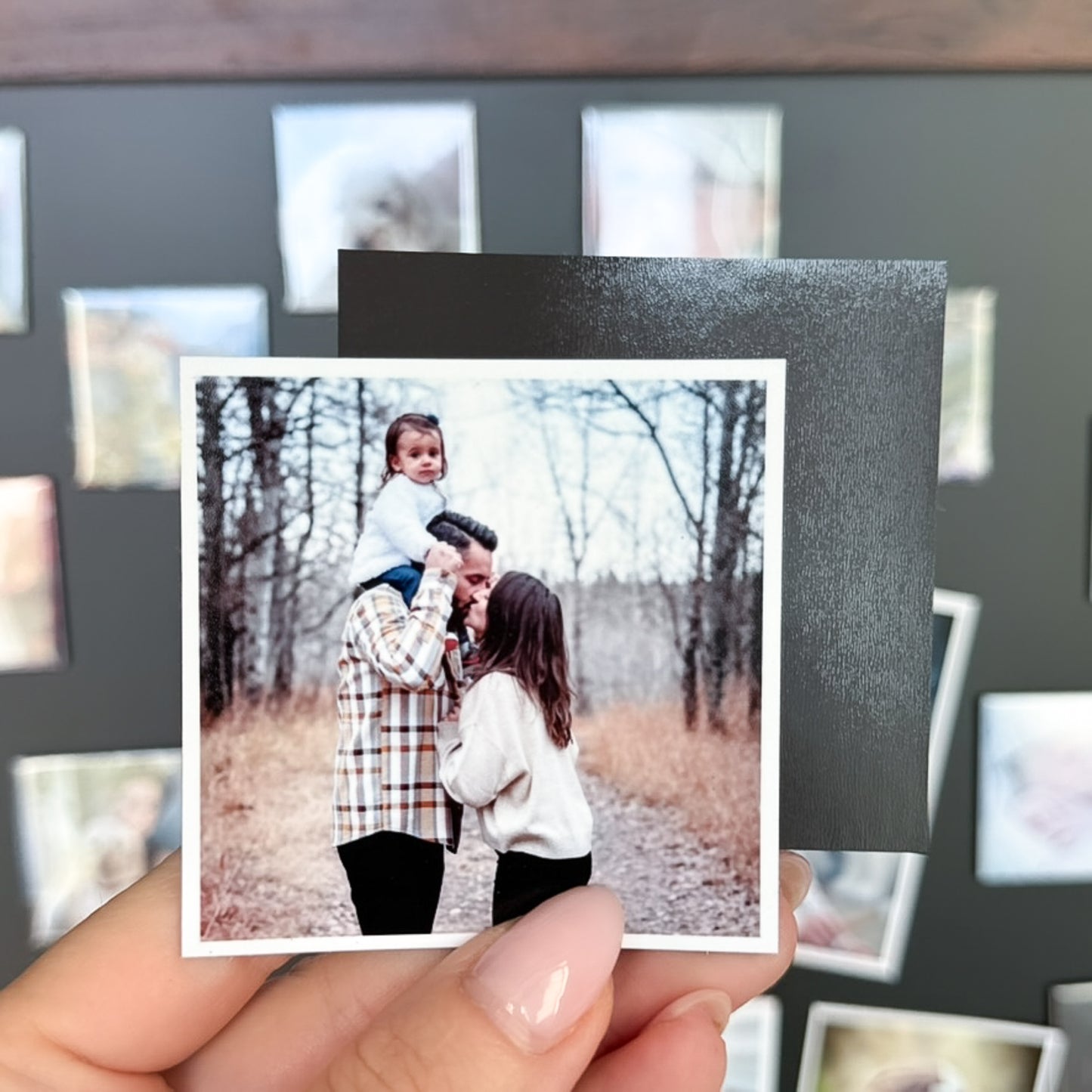 Person holding a photo magnet of a couple with a child outdoors, with a blurred collage of other photo magnets in the background.