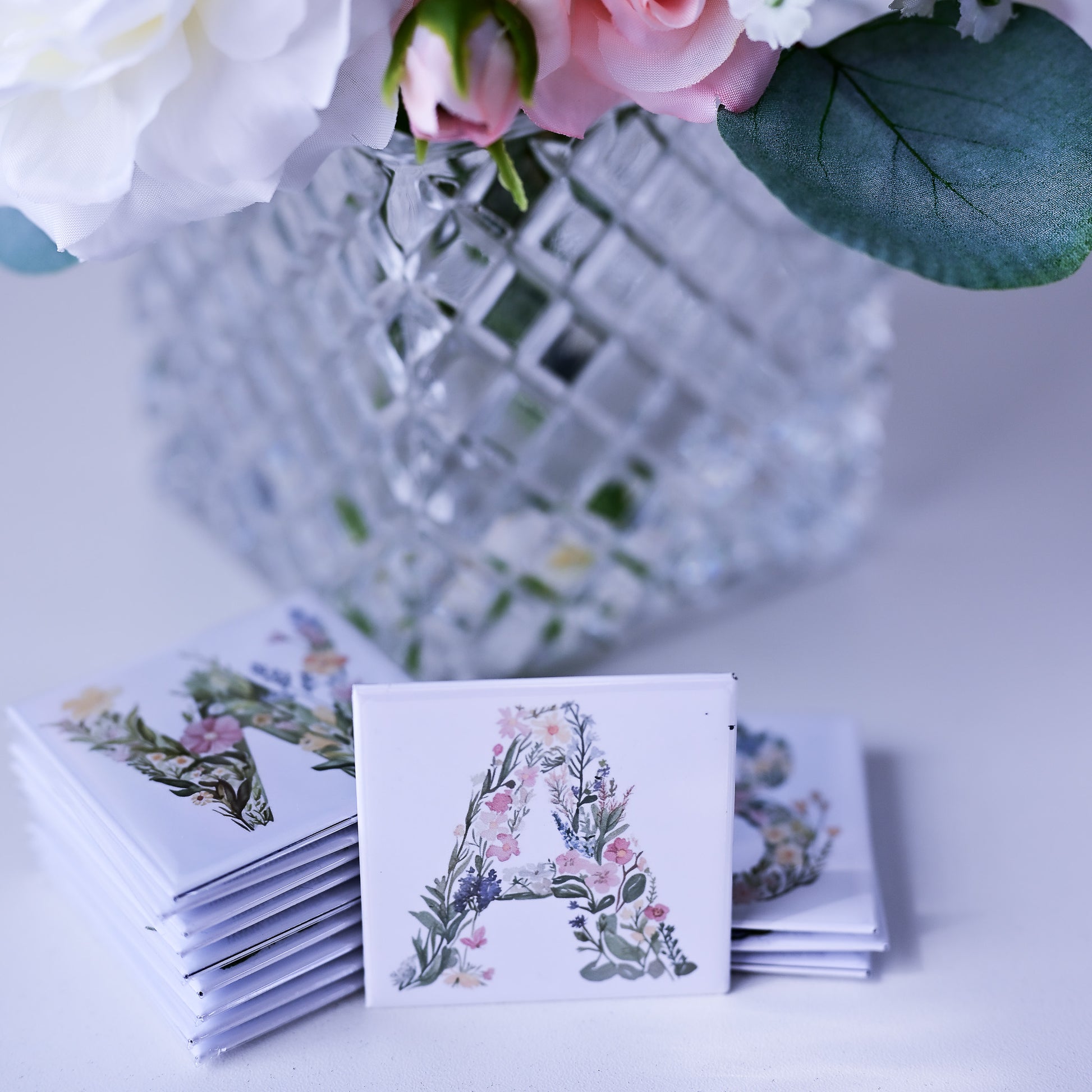 Stack of fridge magnets with floral design and letter 'A' on a white surface with flowers in the background