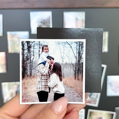 Person holding a photo magnet of a couple with a child outdoors, with a blurred collage of other photo magnets  in the background.